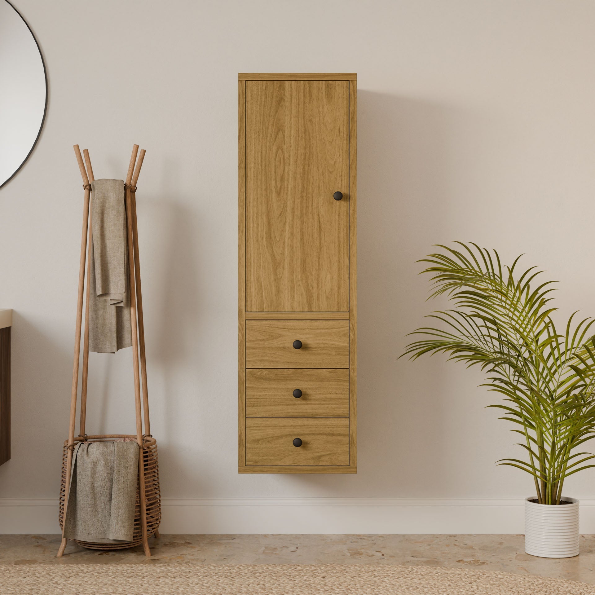 A minimalist interior featuring a Krovel Furniture Co. Triple Drawer Linen Cabinet in White Oak with a tall cupboard and three drawers, a round wall mirror, wooden coat rack with towels, a woven basket below, and a potted palm plant on the light floor.